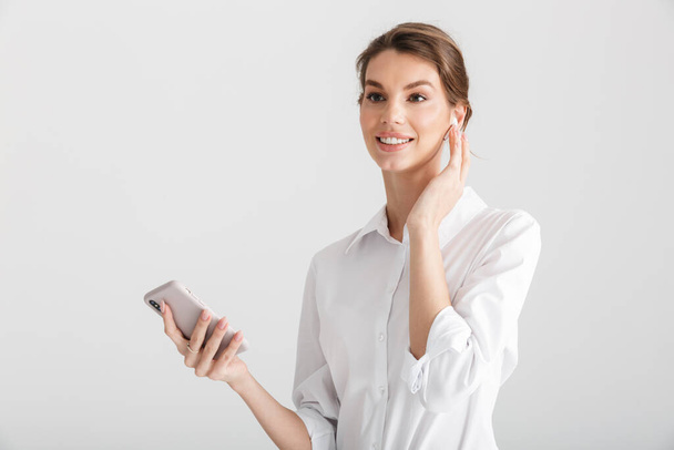 Изображение улыбающейся красивой женщины, использующей беспроводной наушник и мобильный телефон на белом фоне
 - Фото, изображение