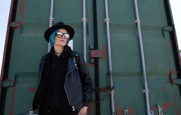Girl with blue hair in a black hat and leather jacket poses near a green container. - Photo, image