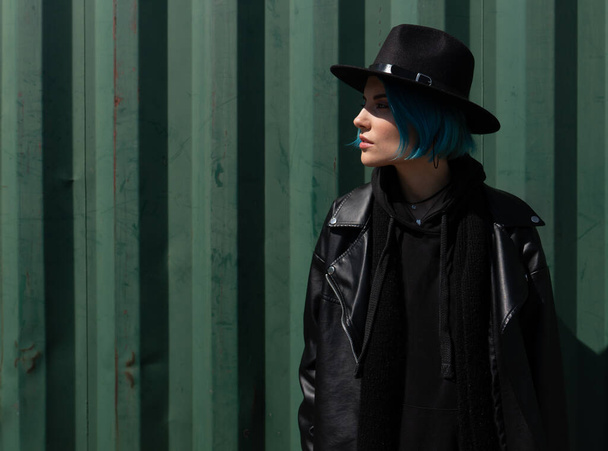 Ragazza con i capelli blu in un cappello nero e giacca di pelle posa vicino a un contenitore verde. Copia spazio - Foto, immagini