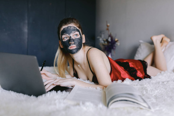 Student with mud mask reading textbook and using laptop - Фото, изображение
