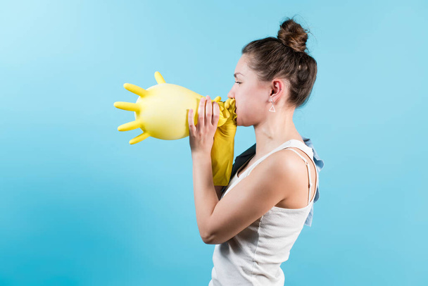 Side view of a young woman in a t-shirt inflates a yellow household glove. Isolated on blue background. - Photo, Image