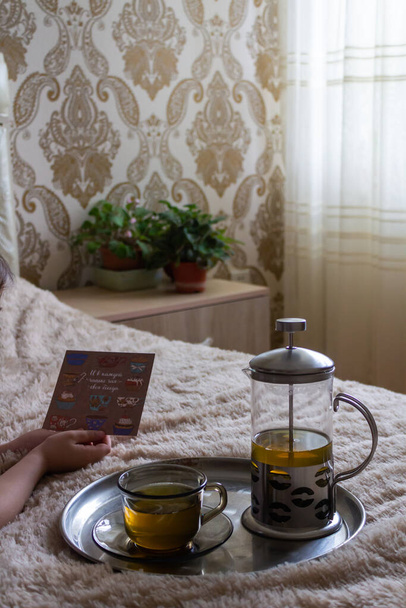 A gyermek az ágyon fekszik, és kezében egy kártyát tart a tálca mellett egy csészével és teáskanna teával. - Fotó, kép
