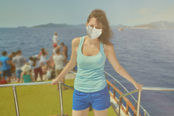 Young beautiful caucasian woman with disposable facial protective medical mask on her face as a protection from coronavirus covid-19 infection is standing on yacht, which is in sea during excursion. - Φωτογραφία, εικόνα
