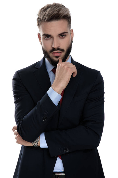 sensual young guy in suit holding arms in a fashion pose and touching chin, standing isolated on white backgrond - Zdjęcie, obraz