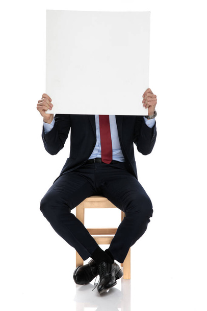 νεαρός επιχειρηματίας κρύβεται πίσω από άδειο πίνακα, κάθεται απομονωμένος σε λευκό φόντο - Φωτογραφία, εικόνα