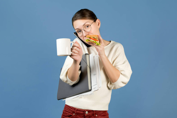 the junior office employee is overworked with work and eats on the go. Girl with a sandwich and coffee on a blue background holds documents and speaks on the phone, looks at the camera - 写真・画像
