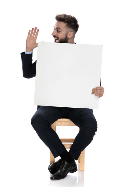 young businessman smiling and holding hands up, holding empty board and saluting, sitting isolated on white background - Photo, Image