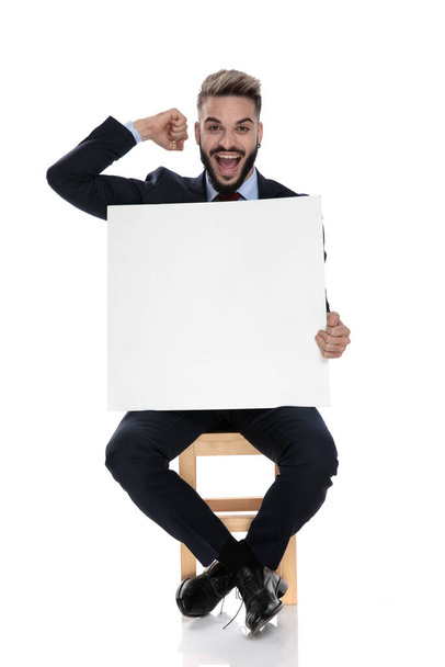 onnellinen nuori liikemies, jolla on tyhjä lauta ja juhlii voittoa, pitämällä nyrkkiä ilmassa, istuen eristyksissä valkoisella taustalla
 - Valokuva, kuva