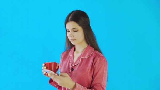 Chica feliz en pijama es beber té o café y el uso de teléfono sobre fondo azul en el estudio
 - Imágenes, Vídeo