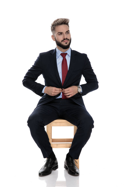 confident young businessman in suit fixing coat, sitting isolated on white background - Fotoğraf, Görsel