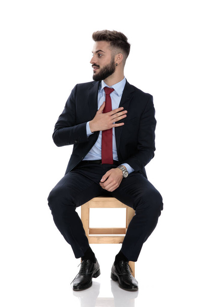 joven hombre de negocios mirando a un lado y sosteniendo la mano en el pecho, sentado aislado sobre fondo blanco
 - Foto, imagen