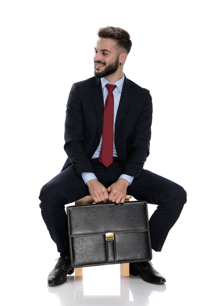 幸せな若いビジネスマンがスーツケースを持って笑顔で白い背景に孤立して座って - 写真・画像