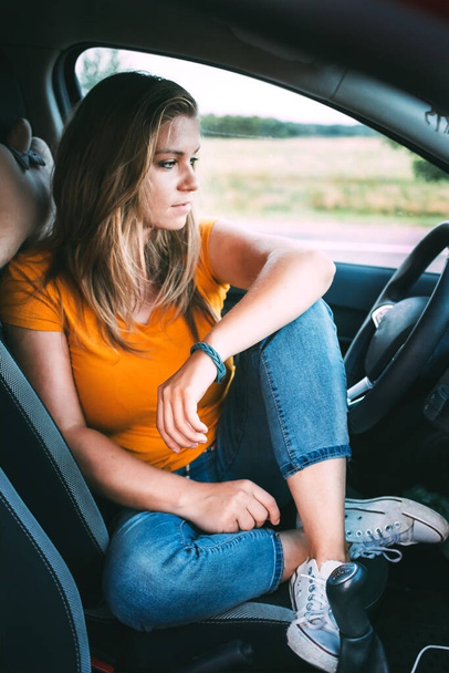  Een blonde vrouw in blauwe jeans en een geel T-shirt zit in een auto voor het stuur en kijkt naar de weg, denkend aan reizen en de weg. Genieten van het rijden - Foto, afbeelding