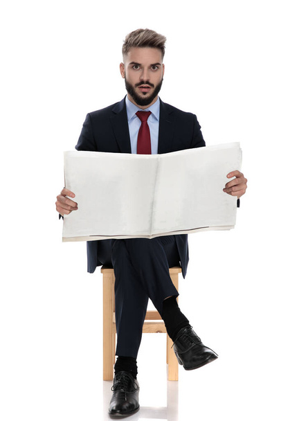 шокований молодий хлопець у костюмі читає газету і сидить ізольовано на білому тлі
 - Фото, зображення