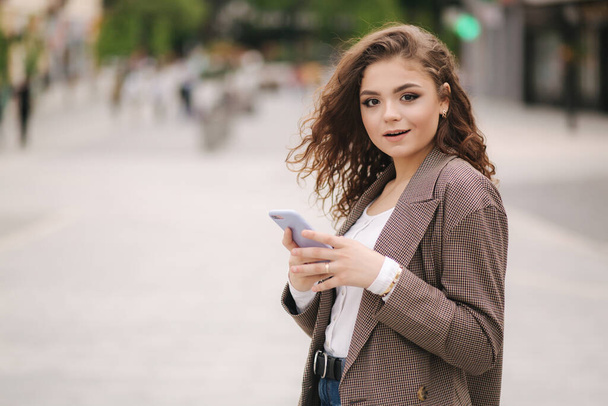 Young woman with curly hair looking in the phone and surprised. Happy female use phone outdoors and amazed - Фото, зображення
