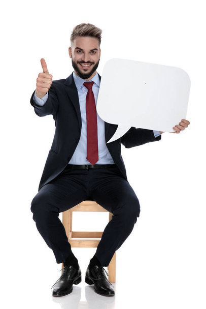 heureux jeune homme d'affaires tenant bulle de discours et faire les pouces vers le haut, assis isolé sur fond blanc
 - Photo, image