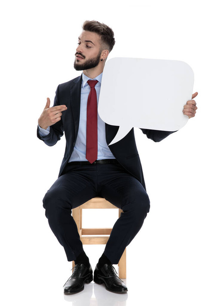 сексуальный молодой бизнесмен указывая пальцами на пузырь речи и глядя в сторону, сидя изолированы на белом фоне
 - Фото, изображение