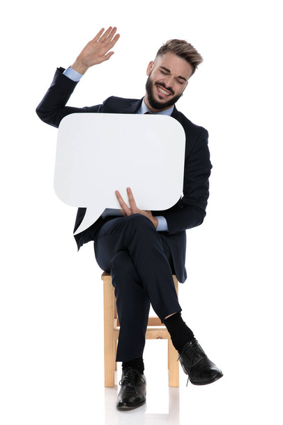happy young businessman holding speech bubble and laughing, sitting isolated on white background - Photo, image