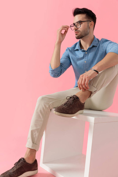 zijaanzicht van een jonge zakenman in blauw shirt zittend met been rustend op kubus zijn hand opheffen tot bril dramatisch op roze studio achtergrond - Foto, afbeelding