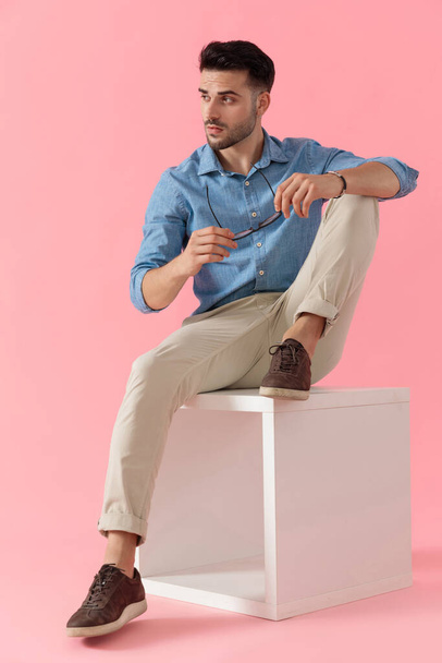 sexy zakenman dragen van blauw shirt zitten met een been rusten op kubus terwijl het nemen van een bril af en kijken opzij cool op roze studio achtergrond - Foto, afbeelding