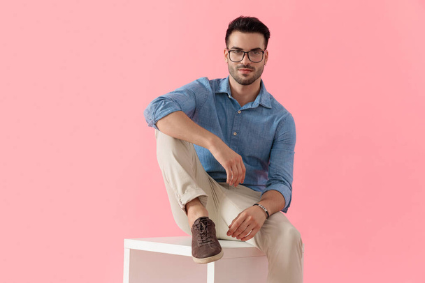 handsome businessman wearing glasses sitting with leg resting on cube and looking at camera happy on pink studio background - Foto, Imagem