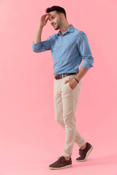 Seitenansicht eines sexy Geschäftsmannes in blauem Hemd, der mit einer Hand in der Tasche geht, während er mit der anderen seine Augen vor Sonnenlicht schützt und vor rosa Studiohintergrund glücklich nach vorne blickt - Foto, Bild