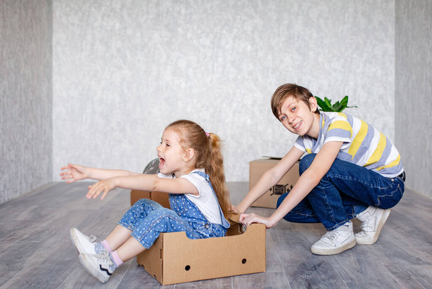 Brother rides sister in a cardboard box. Children play with boxes at home in quarantine. A boy and a girl have moved into a new apartment and are happily playing without toys. - Фото, зображення