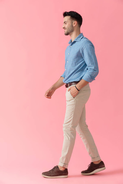 side view of a young businessman wearing blue shirt walking with hand in pocket and looking ahead happy on pink studio background - Photo, image