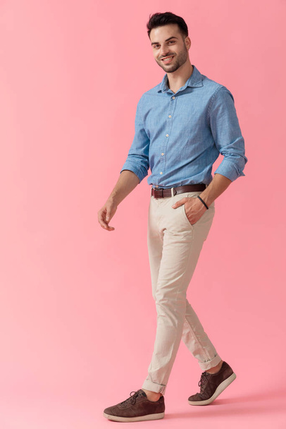 Вид сбоку молодого бизнесмена в голубой рубашке, который ходит с одной рукой в кармане и улыбается на розовом студийном фоне
 - Фото, изображение