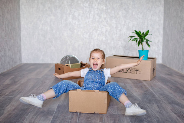A little blonde girl plays at home in quarantine with cardboard boxes. The child moves to a new apartment, is happy and happy. - Foto, afbeelding