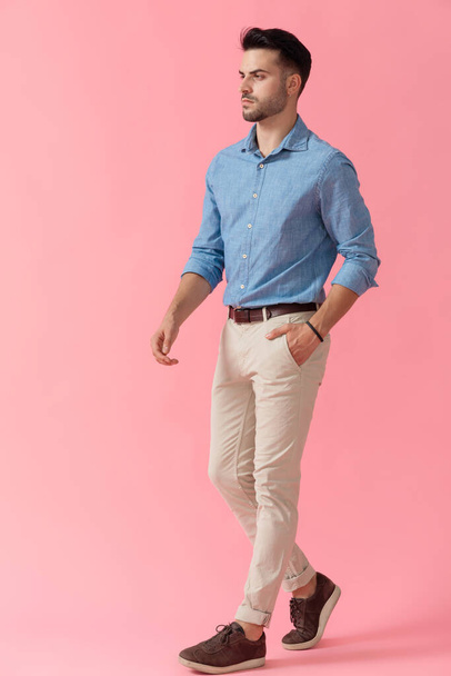 boční pohled na sexy obchodník v modré košili chůze s rukou v kapse a při pohledu dopředu vážné na růžové pozadí studia - Fotografie, Obrázek