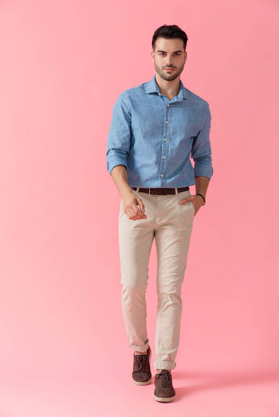 young businessman wearing blue shirt walking with one hand in pocket cool on pink studio background - Photo, image