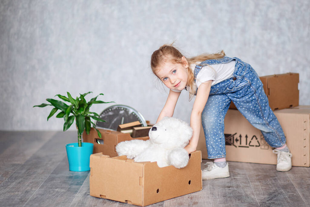A small cheerful blonde girl plays with cardboard boxes and a white Teddy bear in a new house after moving. Unpacking things from cardboard boxes in quarantine. - Photo, image