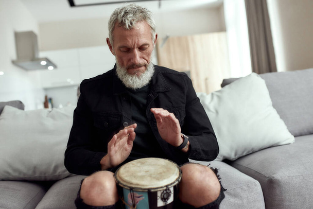Music brings harmony. Bearded middle-aged man, artist or composer writing song, playing rhytm with djembe drum, sitting on the couch at home - Photo, image