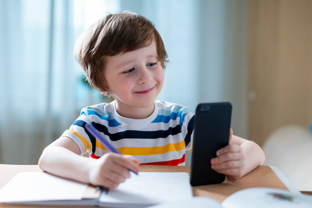 Távolsági tanulás online oktatás. Kaukázusi mosolygós fiú otthon tanul okostelefonnal és házi feladatot csinál. Gondolkodó gyermek ül az asztalnál a mobiltelefon miatt coronavirus karantén. - Fotó, kép