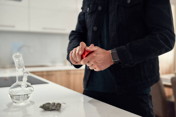 Cropped shot of man using red marijuana grinder, while standing in the kitchen. Glass water pipe or bong, cannabis buds in a plastic bag on the table - Photo, image