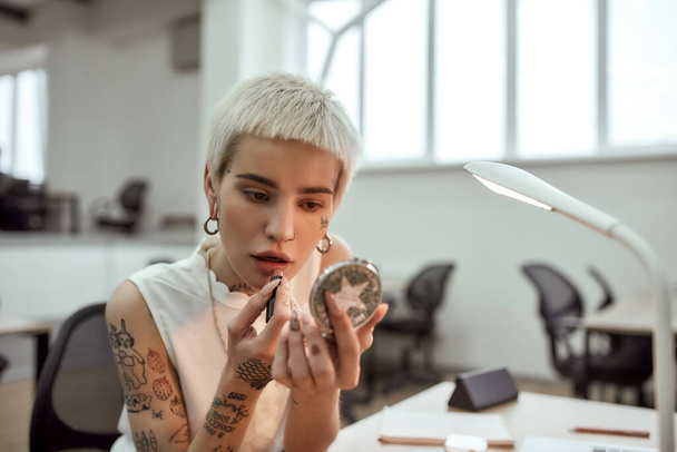 Adjusting her makeup. Portrait of young and stylish blonde tattooed business woman applying lipstick while sitting in the modern coworking space - Φωτογραφία, εικόνα