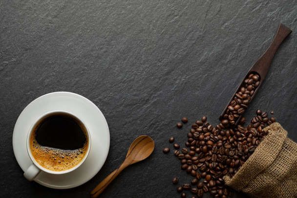 Top view above of Black hot coffee for morning with milk foam for morning menu in white ceramic cup with coffee beans roasted in sack bag on dark stone table background. Flat lay with copy space. - Fotoğraf, Görsel