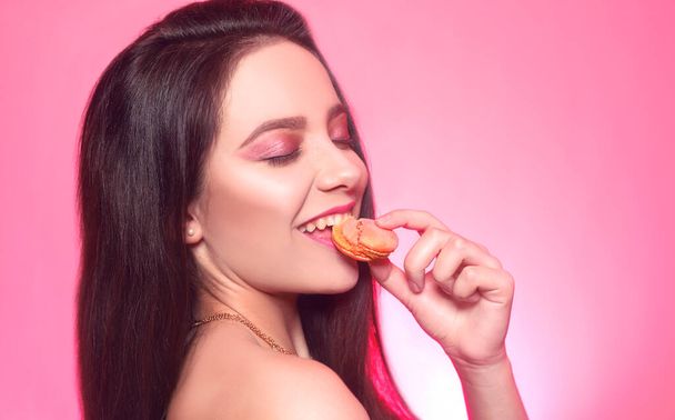 Дівчина їсть макаруни, модель на рожевому фоні їсть солодку, цукрову дієту. Брюнетка з рожевим макіяжем, залежна від їжі. Укуси макарунів, печиво
 - Фото, зображення