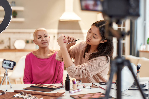 Speak the beauty. Asian female blogger showing lipstick colors while applying makeup on middle aged woman face using cosmetics on the table. Two women recording a tutorial video for beauty blog - Photo, image