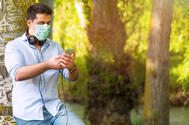 Jonge man met beschermende gezichtsmasker luisteren naar muziek buiten in het park met behoud van sociale afstand. Lifestyle tijdens Covid-19 pandemische uitbraak. - Foto, afbeelding