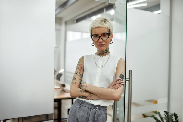 Successful business lady. Young beautiful blonde tattooed businesswoman with short haircut leaning on the glass door, keeping arms crossed and looking at camera while standing in modern office - Photo, image