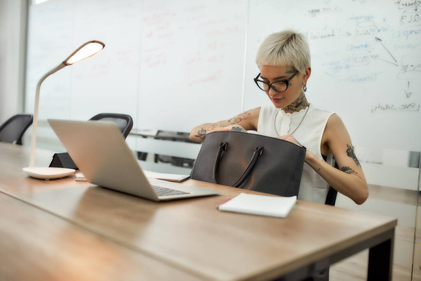Jonge aantrekkelijke blonde getatoeëerde zakenvrouw met kort kapsel op zoek naar iets in haar zwarte handtas tijdens het werken in het moderne kantoor - Foto, afbeelding