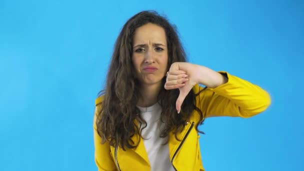Retrato de mujer seria con chaqueta amarilla mostrando pulgares hacia abajo signo de disgusto en el estudio con fondo azul
. - Imágenes, Vídeo
