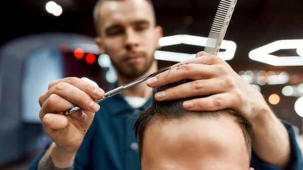 Personal stylist. Close up of young professional barber with scissors and hair comb in hands doing haircut for his client in barber shop - Foto, afbeelding