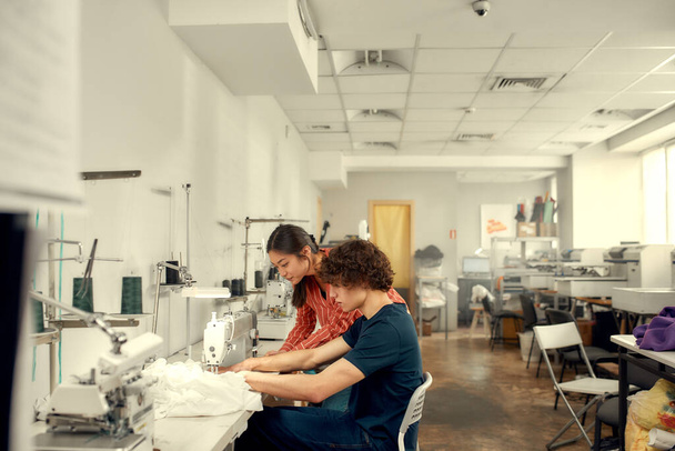 Careful measures. Young man, clothing designer sitting and sewing. His female colleague helping him. Dressmaker working on the sewing machine. Tailor making a garment in his workplace - Foto, Bild