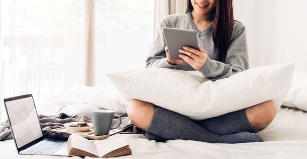 Genç Asyalı kadın evdeki yatakta dijital tablet bilgisayar kullanıyor ve rahatlıyor. Sosyal uygulamaları kontrol ediyor ve oyun oynuyor. İletişim ve teknoloji kavramı. - Fotoğraf, Görsel