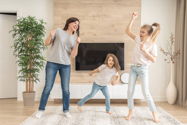 Mutlu anne ve iki kız saç fırçasıyla karaoke şarkı söyleyerek eğleniyorlar. Anne gülerek evde genç bir kızla eğlenceli bir yaşam tarzı aktivitesi yapıyor.. - Fotoğraf, Görsel