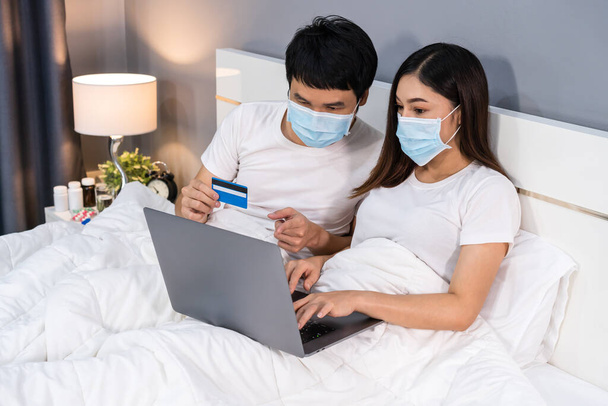 Pari lääketieteelliset naamiot kannettavan tietokoneen avulla ostoksia verkossa luottokortilla sängyllä aikana karanteeni pandemia coronavirus (covid-19) pysyä kotona. - Valokuva, kuva