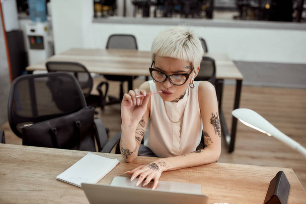 Full concentration. Serious young blonde tattooed businesswiman in wireless earphones sitting at her working place, looking at the screen of laptop and thinking about something with pen in her mouth - Foto, imagen